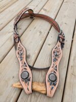 A tan leather horse bridle with intricate cutout designs and silver floral accents lies on a wooden surface. The bridle includes adjustable buckles and detailed tooling, with teal highlights accentuating the patterns.