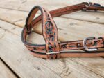 A close-up of a brown leather belt with intricate teal and black patterns engraved on it. The belt is placed on a wooden surface with visible wood grain. The belt has metal buckles and multiple holes for size adjustment.