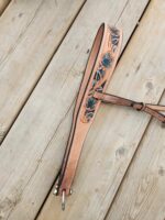 A leather belt with intricate blue floral embroidery lies on a wooden deck. The stitching is done in white, and the belt features a silver buckle. The wood planks underneath have a natural finish with visible grain and knots.