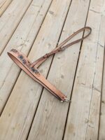 A tan leather guitar strap with floral and leaf patterns is laid out on a wooden deck. The strap includes metal buckles for adjusting the length. The wood boards of the deck have visible grain and knots.