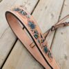 Close-up of a leather belt with intricate tooled floral designs and blue accents, laid on a wooden surface. The belt's craftsmanship and detail are highlighted, showcasing the delicate patterns and stitching.