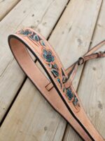 Close-up of a leather belt with intricate tooled floral designs and blue accents, laid on a wooden surface. The belt's craftsmanship and detail are highlighted, showcasing the delicate patterns and stitching.