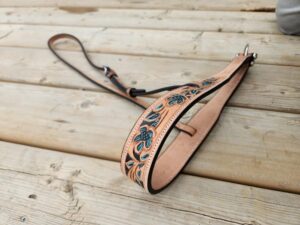 A decorative leather strap with intricate floral patterns in blue and green is laid out on a wooden surface. The strap appears handcrafted and features metal hardware at both ends. The wood has a rustic, weathered look with visible grain and knots.