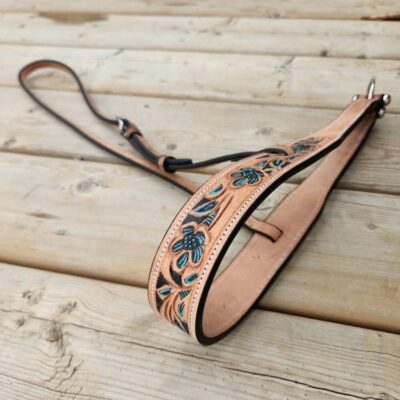 A decorative leather strap with intricate floral patterns in blue and green is laid out on a wooden surface. The strap appears handcrafted and features metal hardware at both ends. The wood has a rustic, weathered look with visible grain and knots.