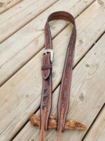 A brown leather belt with decorative turquoise cutouts and a silver buckle is looped on a wooden plank. The leather has intricate stitching along its edges and the belt is secured with a small leather strap. The wooden surface appears weathered and rustic.