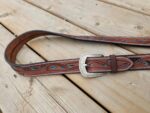 A brown leather belt with intricate turquoise and black leaf-like patterns is laid out on a wooden surface. The belt features white stitching along the edges and has a detailed metal buckle with engraved designs.