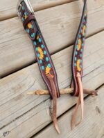 A pair of decorated leather straps with colorful floral embroidery and turquoise accents are laid out on a wooden surface. The straps have intricate patterns and are designed with buckles for fastening, with loose leather ties at the ends.