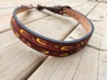 A handcrafted leather belt with a brown and black design, featuring intricate red and yellow feather patterns. The belt is lying on a wooden surface, with the buckle end slightly blurred in the background.