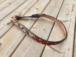 A leather belt with colorful floral embroidery, featuring yellow, orange, and blue patterns, is displayed on a wooden surface. The belt has a dark brown outer edge with a metal buckle and tan-colored inner lining.