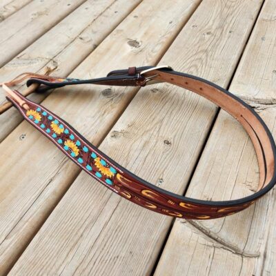 A leather belt with colorful floral embroidery, featuring yellow, orange, and blue patterns, is displayed on a wooden surface. The belt has a dark brown outer edge with a metal buckle and tan-colored inner lining.