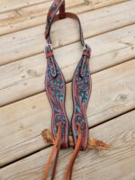 A decorative leather horse bridle with intricate floral patterns in turquoise and brown lies on a wooden surface. Two tan leather straps hang from the bottom, and the bridle features detailed stitching and silver hardware.