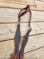 A close-up of a decorated leather horse bridle on a wooden surface. The bridle features intricate teal and black floral designs with silver buckles and hardware. The background consists of weathered wooden planks.