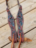 A pair of brown leather reins with intricate teal embroidery and floral patterns lies on a wooden surface. The reins have metal buckles and tan leather straps at the ends, showcasing a mix of traditional craftsmanship and detailed artistry.