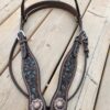 A detailed brown leather horse bridle decorated with elaborate turquoise stitching and silver conchos lies on a wooden deck. The conchos feature intricate patterns, and the bridle includes silver buckles and straps. A hand is visible holding one end of the bridle.