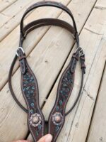 A detailed brown leather horse bridle decorated with elaborate turquoise stitching and silver conchos lies on a wooden deck. The conchos feature intricate patterns, and the bridle includes silver buckles and straps. A hand is visible holding one end of the bridle.