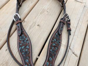 A detailed brown leather horse bridle decorated with elaborate turquoise stitching and silver conchos lies on a wooden deck. The conchos feature intricate patterns, and the bridle includes silver buckles and straps. A hand is visible holding one end of the bridle.