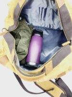 An open tan and brown The Rocky Mountain Perfect Duffel- Explorer Series showing its contents, which include a green blanket or jacket and a purple water bottle. The interior lining of the bag is gray, with pockets visible inside, making it perfect for carrying your Rocky Mountain hiking gear on outdoor adventures.