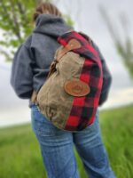 A person wearing a gray hoodie and blue jeans is standing outdoors with their back to the camera. They are carrying The Rocky Mountain Sling Bag- Explorer Series, which is made from wax canvas, half olive green and half red-and-black plaid, with a rectangular leather patch on the side. The background is grassy and slightly blurred.