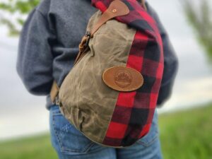 A person wearing a gray hoodie and blue jeans is standing outdoors with their back to the camera. They are carrying The Rocky Mountain Sling Bag- Explorer Series, which is made from wax canvas, half olive green and half red-and-black plaid, with a rectangular leather patch on the side. The background is grassy and slightly blurred.