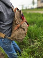 A person wearing jeans and a grey hoodie is carrying The Rocky Mountain Sling Bag- Explorer Series with leather straps over one shoulder while standing on a grassy area. The bag has a glimpse of red and black plaid fabric. The background is blurred, with a hint of a building, evoking the spirit of Rocky Mountain hiking gear.