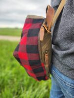 A person in a gray sweater and blue jeans carries The Rocky Mountain Sling Bag- Explorer Series while standing in a grassy field under an overcast sky.