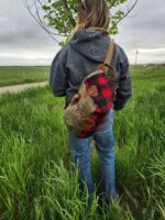 A person in a gray hoodie and blue jeans stands in a grassy field, facing away from the camera. They have The Rocky Mountain Sling Bag- Explorer Series slung over one shoulder. A small tree and an overcast sky create a serene backdrop, reminiscent of Rocky Mountain trails.