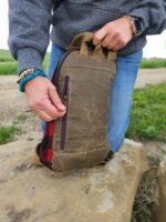 A person wearing a gray sweater and blue jeans is unzipping The Rocky Mountain Sling Bag- Explorer Series. The individual has bracelets on their wrist and an electronic watch. The scene appears to be outdoors, with green grass and a dirt path in the background, possibly in the Rocky Mountains.