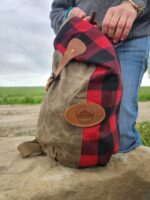 Close-up of a person holding The Rocky Mountain Sling Bag- Explorer Series, made of brown wax canvas and red-and-black plaid fabric, complete with a leather patch. The person's hand, adorned with pink nail polish, ties this scene together against the backdrop of the Rocky Mountain hiking gear setting with grass and a dirt path.