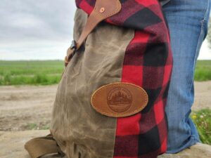 Close-up of a person holding The Rocky Mountain Sling Bag- Explorer Series, made of brown wax canvas and red-and-black plaid fabric, complete with a leather patch. The person's hand, adorned with pink nail polish, ties this scene together against the backdrop of the Rocky Mountain hiking gear setting with grass and a dirt path.