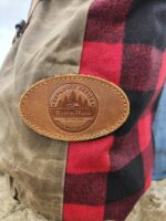 Close-up of a zip up leather backpack with red and black plaid fabric and a brown leather patch. The patch has an embossed design with mountains, trees, and text that reads "The Rocky Mountain Sling Bag- Explorer Series, Ranch Hand, High Mountain Tough." The waxed canvas fabric beside it is olive green.