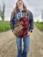 A person with long hair stands on a gravel pathway, wearing a gray hoodie with a logo, jeans, and holding The Rocky Mountain Sling Bag- Explorer Series. The individual is looking upwards with a broad smile. In the background, there is greenery and a cloudy sky.