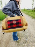 A person in a grey jacket and jeans stands on a dirt path holding a yellow duffle bag with a red and black plaid pattern and a circular brown leather patch. The path, perfect for The Rocky Mountain Sling Bag- Explorer Series, is surrounded by green grass and trees.