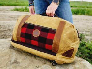 Person with a zip-up grey sweatshirt and blue jeans holding The Rocky Mountain Sling Bag- Explorer Series, featuring a red and black checkered pattern and brown leather accents. The bag, reminiscent of rugged Rocky Mountain hiking gear, is resting on a large rock, with a rural landscape featuring a dirt road and grassy fields in the background.