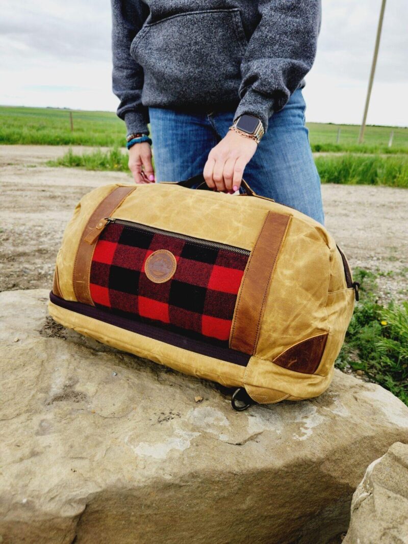 Person with a zip-up grey sweatshirt and blue jeans holding The Rocky Mountain Sling Bag- Explorer Series, featuring a red and black checkered pattern and brown leather accents. The bag, reminiscent of rugged Rocky Mountain hiking gear, is resting on a large rock, with a rural landscape featuring a dirt road and grassy fields in the background.