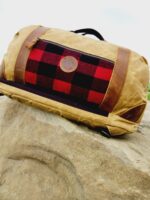 A tan duffel bag with brown leather accents and a red-and-black plaid pocket is placed on a large rock. The Rocky Mountain Sling Bag- Explorer Series, perfect for rocky mountain hiking gear, has a circular leather logo patch on the front pocket and black handles. The background features a cloudy sky.