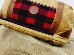 A tan duffel bag with brown leather accents and a red-and-black plaid pocket is placed on a large rock. The Rocky Mountain Sling Bag- Explorer Series, perfect for rocky mountain hiking gear, has a circular leather logo patch on the front pocket and black handles. The background features a cloudy sky.