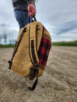 A person wearing blue jeans holds The Rocky Mountain Sling Bag- Explorer Series by its top handle while standing on a gravel path. The sky is cloudy, and the background features blurred greenery and buildings, capturing an essence of Rocky Mountain hiking gear.