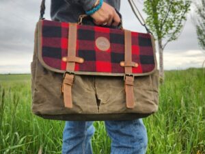 A person standing on grass, holding The Rocky Mountain Sling Bag- Explorer Series with a red and black plaid flap and brown leather straps. The person is wearing blue jeans, a grey sweater, and a blue bracelet. In the background, some trees appear against a cloudy sky—perfect weather for testing Rocky Mountain hiking gear.