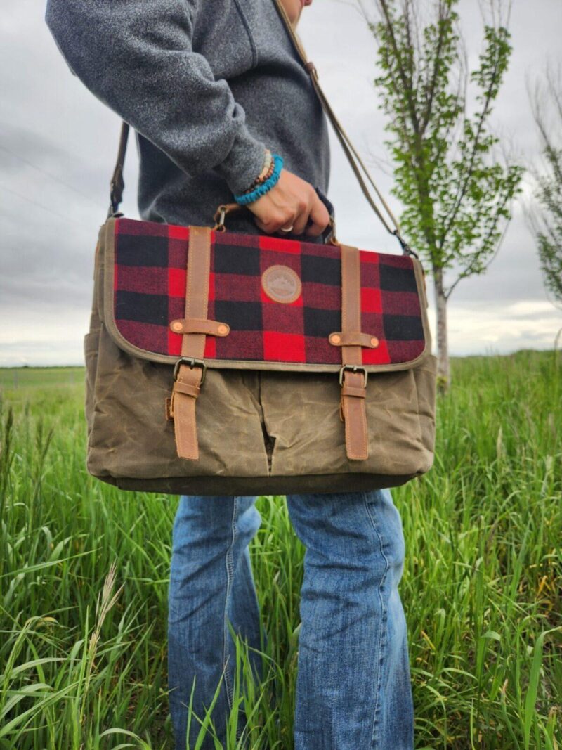 A person standing on grass, holding The Rocky Mountain Sling Bag- Explorer Series with a red and black plaid flap and brown leather straps. The person is wearing blue jeans, a grey sweater, and a blue bracelet. In the background, some trees appear against a cloudy sky—perfect weather for testing Rocky Mountain hiking gear.