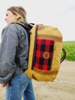 A person wearing a gray jacket and blue jeans is carrying The Rocky Mountain Sling Bag- Explorer Series down a dirt road. The background features a grassy field under a cloudy sky. The person, seemingly equipped with essential hiking gear, is looking off into the distance.