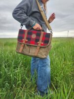 A person stands in a grassy field, holding The Rocky Mountain Sling Bag- Explorer Series with a red and black buffalo plaid flap and brown leather straps. They wear a grey jacket and blue jeans, looking ready for their next Rocky Mountain adventure. The sky is overcast.