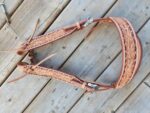 A detailed, intricately carved White Buckstitch Browband Headstall with floral patterns lies on a wooden surface. The headstall, resembling a belt-style headstall, is decorated with ornate silver buckles and leather straps. The wood beneath it has a natural, rustic appearance.