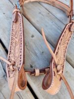 A close-up of a detailed White Buckstitch Browband Headstall with intricate floral patterns, displayed on a wooden surface. The craftsmanship includes embossed designs and neatly stitched edges. Loose straps hang, indicating the headstall is not in use.