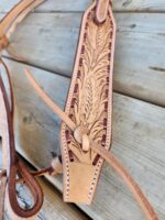 A close-up of a handcrafted leather strap with intricate embossed detailing on a wooden surface. The White Buckstitch Browband Headstall, reminiscent of a belt-style headstall, features leaf-like patterns and is secured with a leather lace threaded through a loop. The craftsmanship highlights fine stitching and carving.