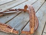 A detailed photo of a White Buckstitch Browband Headstall with intricate floral carvings and silver buckles, resting on a wooden surface. The craftsmanship highlights the fine tooling and stitching in the leather pieces.