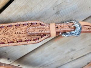 Close-up of a White Buckstitch Browband Headstall with intricate leaf pattern carving and a decorative silver buckle with engraved details, showcasing a refined belt style. The background is a wooden surface with a slightly weathered appearance.