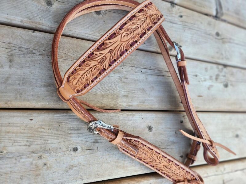 A detailed view of a handcrafted White Buckstitch Browband Headstall with intricate floral carvings. Reminiscent of a belt style headstall, the headstall is placed on a wooden surface, showcasing its rich brown color and fine craftsmanship. The metal buckles add a touch of contrast.