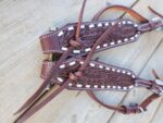 A close-up of intricately designed brown leather straps on a wooden surface. The White Buckstitch Browband Headstall features detailed carved leaf patterns, light-colored edging, and adjustable buckles adorned with silver hardware.