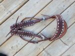 A detailed White Buckstitch Browband Headstall with intricate stitching and white decorative elements is laid out on a wooden surface. The headstall, designed in a belt style, features ornate silver buckles and matching reins.