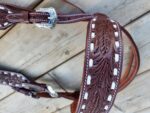 A detailed close-up of a White Buckstitch Browband Headstall with intricate tooled patterns and white stitching. The leather features elaborate leaf designs and silver buckles, showcasing the artistry involved. The background consists of wooden planks, possibly a stable floor.